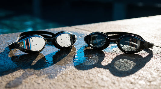 Close-up of mirrored swim goggles next to smoke tinted swim goggles on a wet deck
