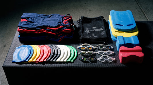 Full team gear kit laid out on a pool deck with suits, caps, goggles, kickboards and mesh bags