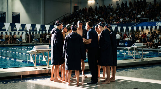 Swim team huddled with their coach on deck at a championship meet in matching warm-up parkas