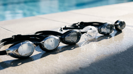 Row of competition swim goggles with mirrored, smoke and clear lenses on a wet pool deck