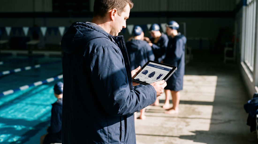 Swim coach on the pool deck using a tablet to set up an online team store, with swimmers in matching team caps and parkas behind