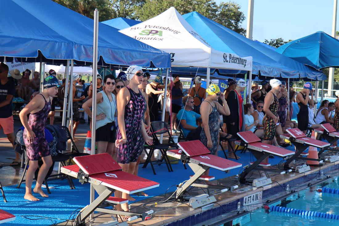 Swimmers and coaches at a poolside event with blue tents and pool equipment.