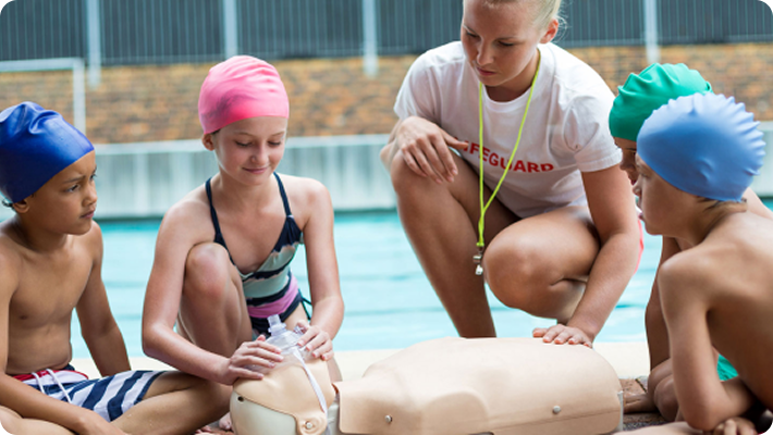 Children learning CPR on a mannequin with a instructor by a pool.